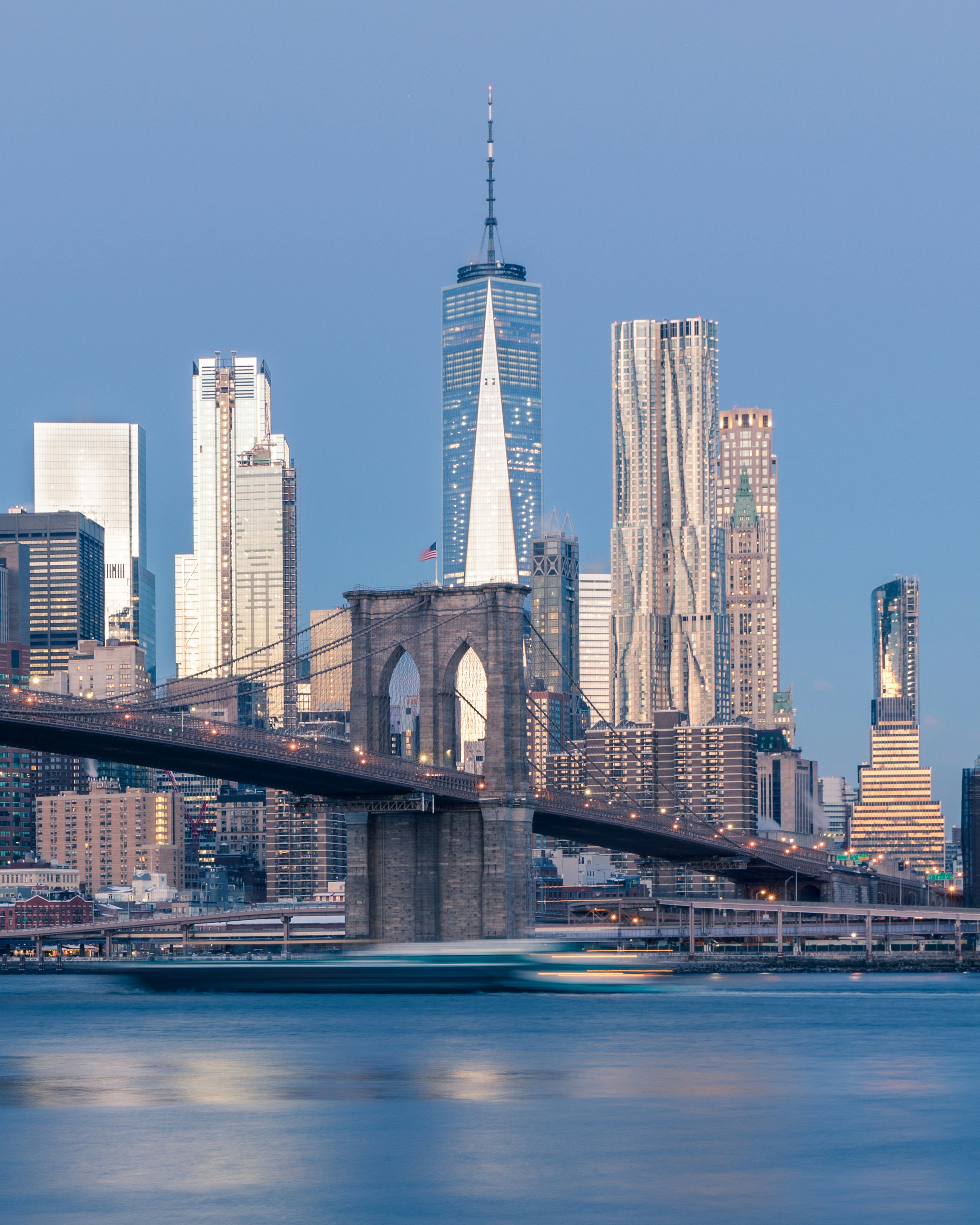 Brooklyn Bridge at dusk with One World Trade Center's spire rising behind it, Lower Manhattan's Financial District skyline reflected in the East River.