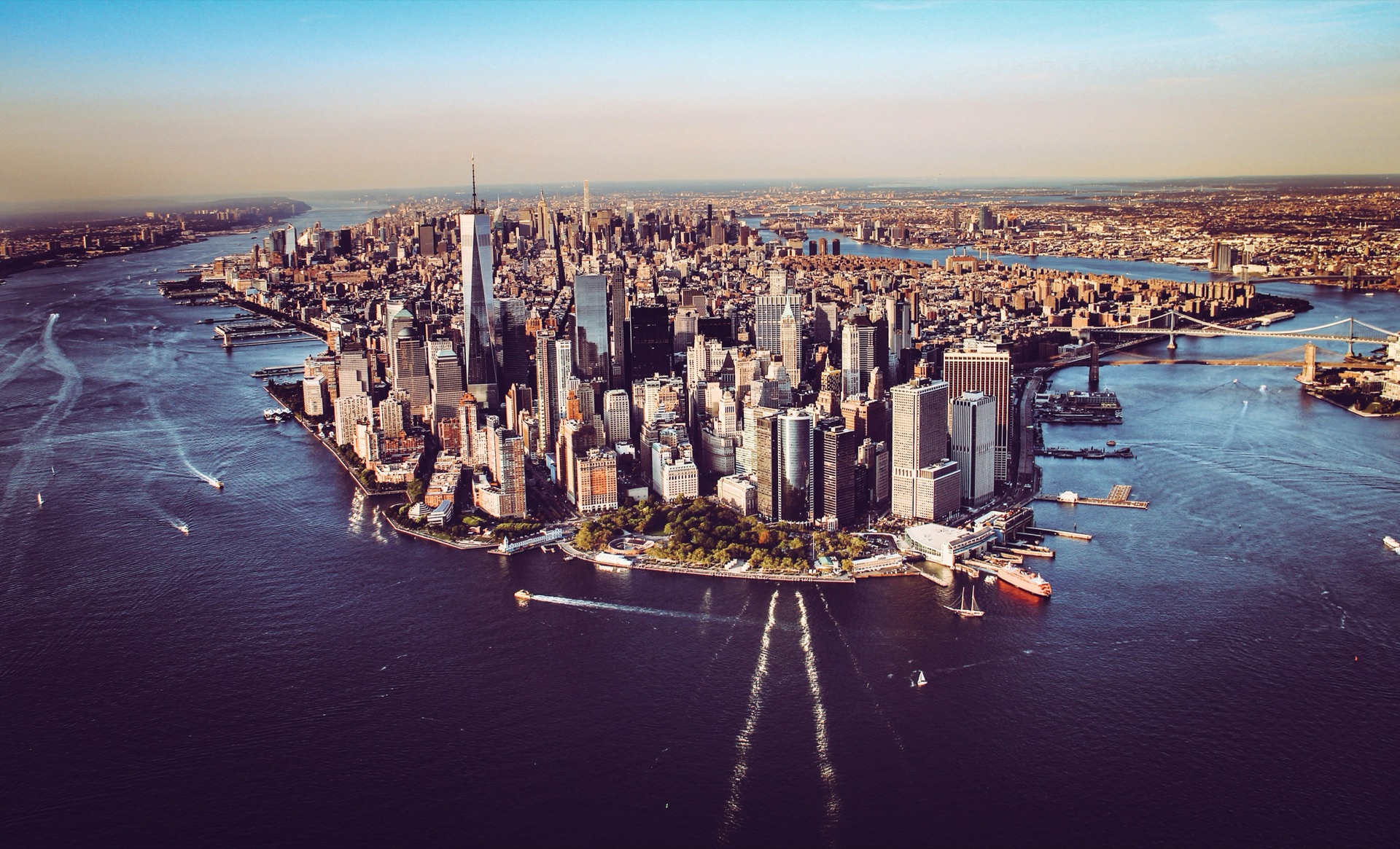 Aerial view of Lower Manhattan at golden hour, One World Trade Center and the Financial District skyline surrounded by the Hudson and East Rivers, with the Brooklyn Bridge visible in the distance.