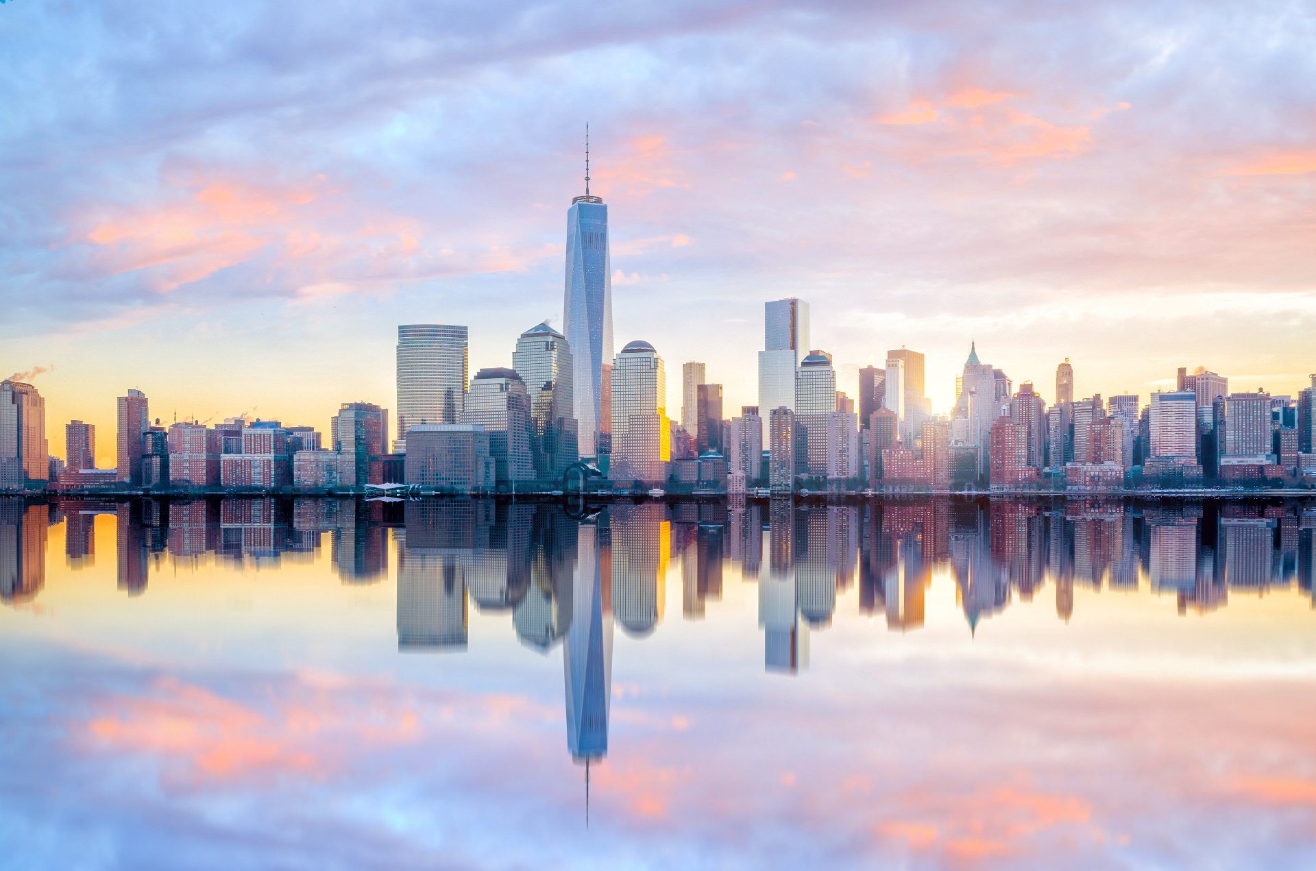 Lower Manhattan skyline at sunrise, One World Trade Center rising above the financial district towers, mirrored in the still water of the harbor under a pink and gold sky.