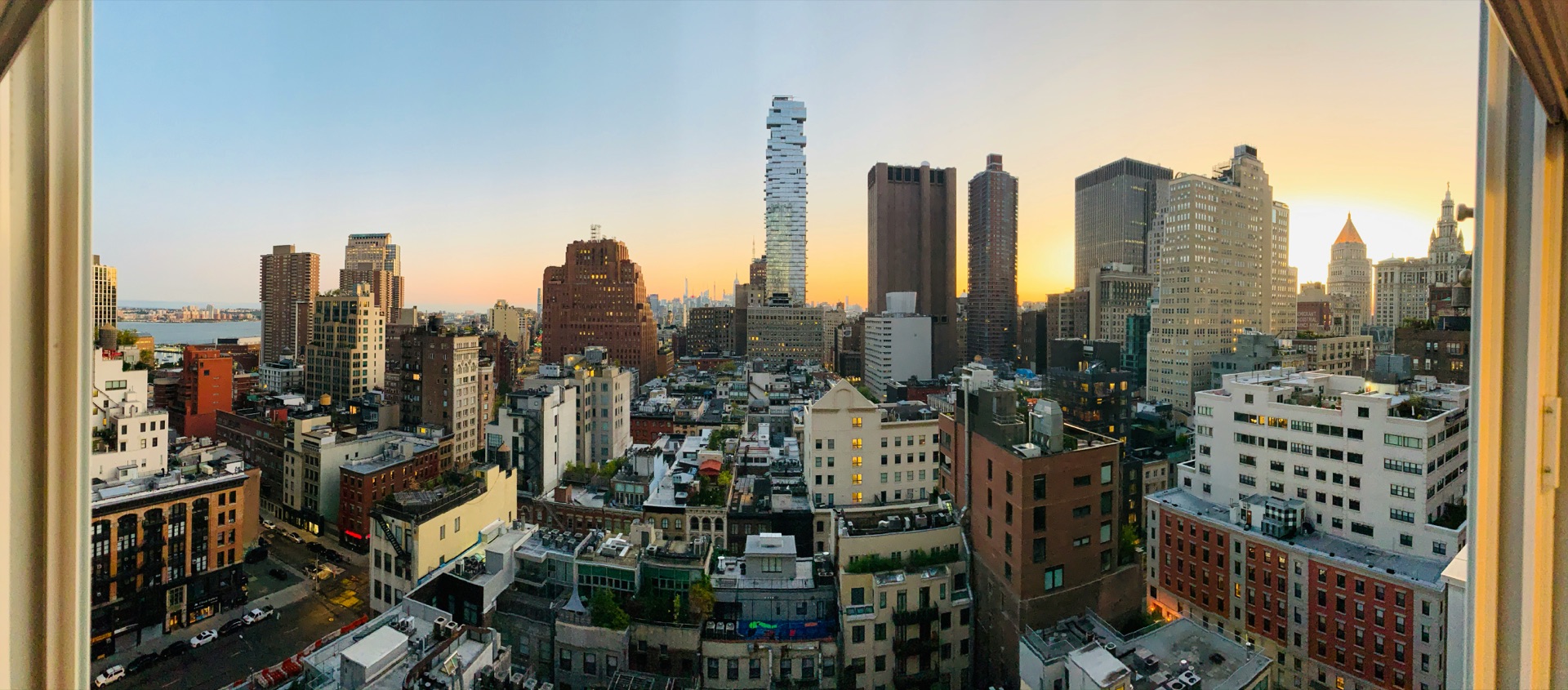 Manhattan skyline at sunset viewed through an open window, downtown towers including 56 Leonard Street rising above dense rooftops, with the Hudson River and New Jersey skyline in the distance.